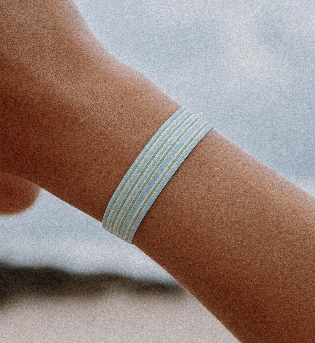 Detailaufnahme eines elastischen Stoffarmbands mit hellblau-grünem Streifenmuster am Handgelenk vor verschwommenem Strandhintergrund.