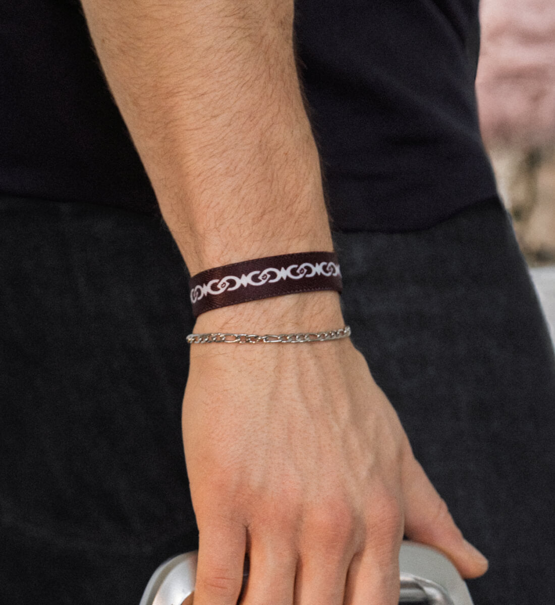Ein schmales, dunkelbraunes Stoffarmband an einem Unterarm, bedruckt mit einem fortlaufenden, weiß-grauen Muster aus verschlungenen, abstrakten Gliedern.