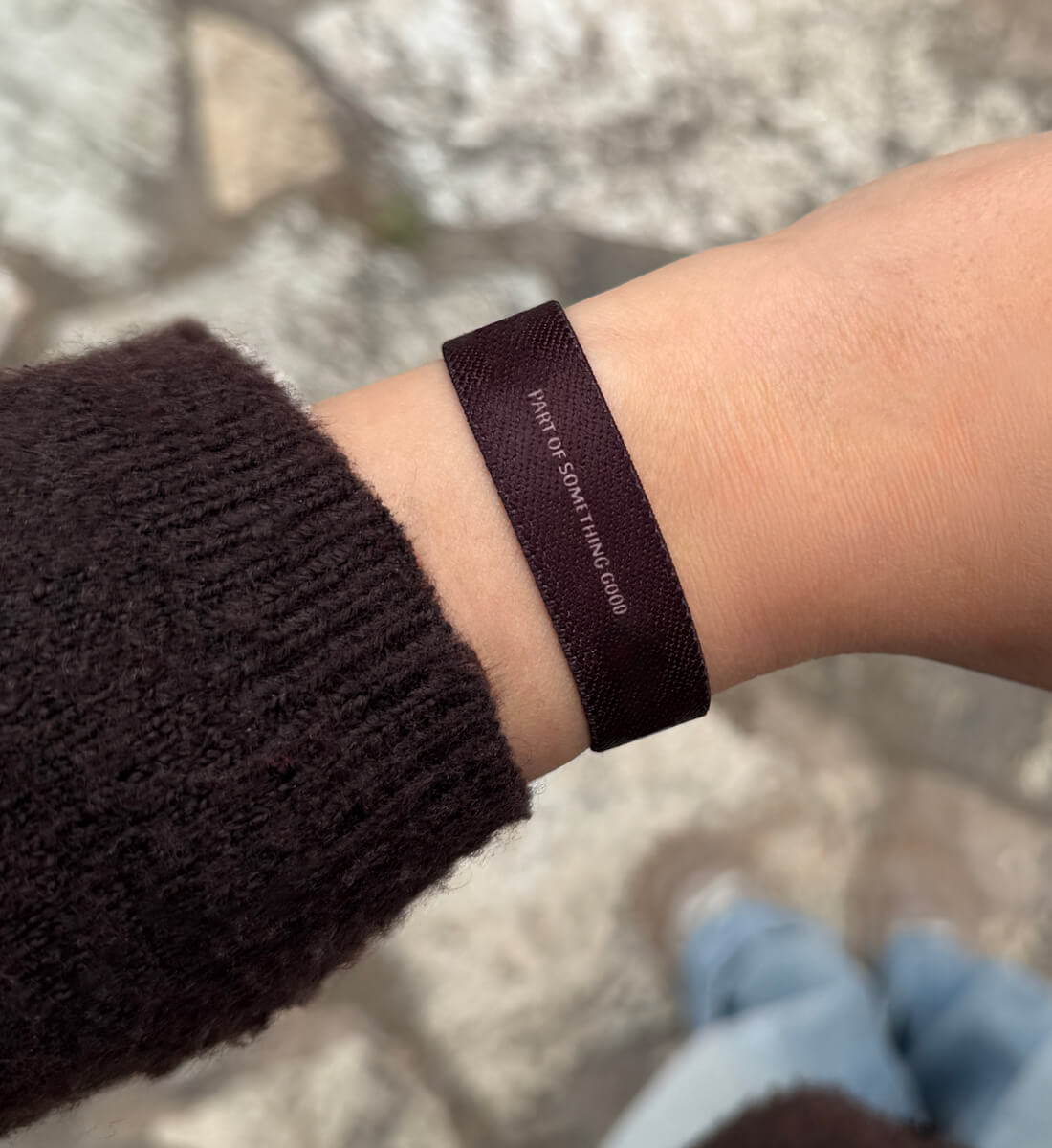 Ein schmales, dunkelbraunes  Armband an einem Handgelenk, auf dem in feiner weißer Schrift der englische Slogan „PART OF SOMETHING GOOD“ steht.