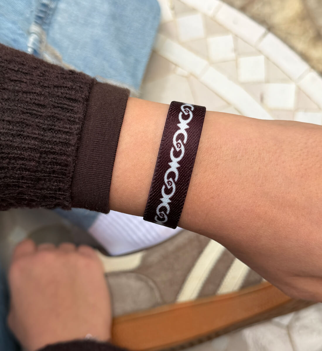 Ein schmales, dunkelbraunes Textilarmband am Handgelenk mit einem kontrastreichen, weißem Muster aus ineinander verschlungenen, geschwungenen Ornamenten.