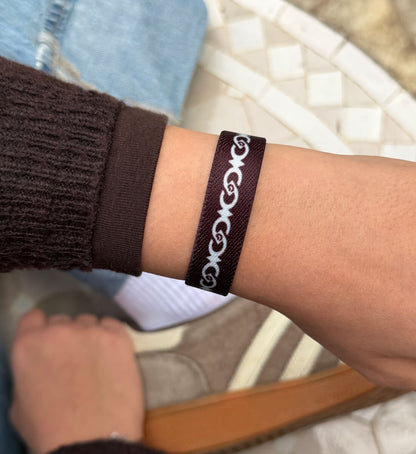 Ein schmales, dunkelbraunes Textilarmband am Handgelenk mit einem kontrastreichen, weißem Muster aus ineinander verschlungenen, geschwungenen Ornamenten.