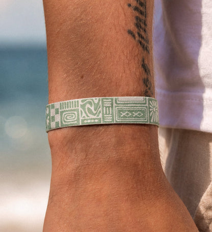 Nahaufnahme eines flach anliegenden, mintgrün gemusterten Stoffarmbands am Handgelenk eines Mannes am Strand.
