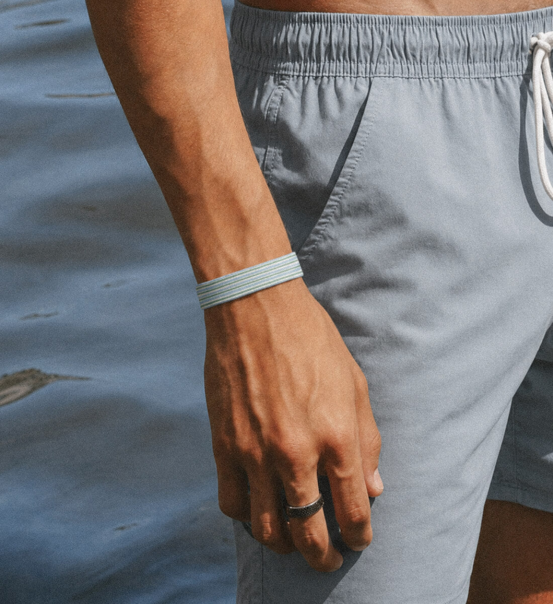 Nahaufnahme eines dünnen, blau-grün gestreiften Stoffarmbands am Handgelenk eines Mannes am Wasser.